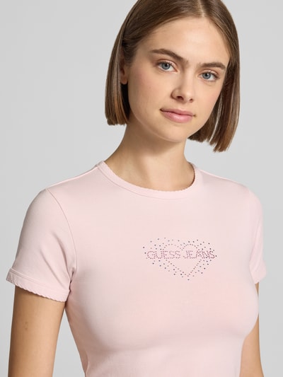 Frauenmodel trägt Slim Fit T-Shirt mit Strasssteinbesatz Modell 'Heart Baby' von GUESS Jeans, Logo-Detail, Rundhalsausschnitt, Nahaufnahme Oberkörper.