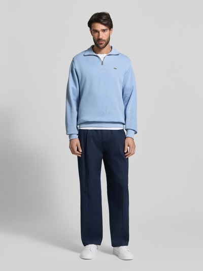 Männermodel trägt Regular Fit Troyer aus reiner Baumwolle von Lacoste in Hellblau, unifarben, mit Logo-Patch und kurzem Reißverschluss, Stricktextur, Ganzkörperansicht von vorne.