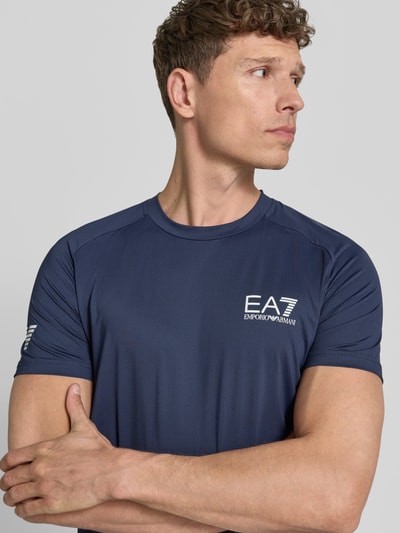 Männermodel trägt T-Shirt mit Label-Print von EA7 Emporio Armani in Dunkelblau, Material-Mix mit Stretch-Anteil, Slim Fit, Nahaufnahme Oberkörper.