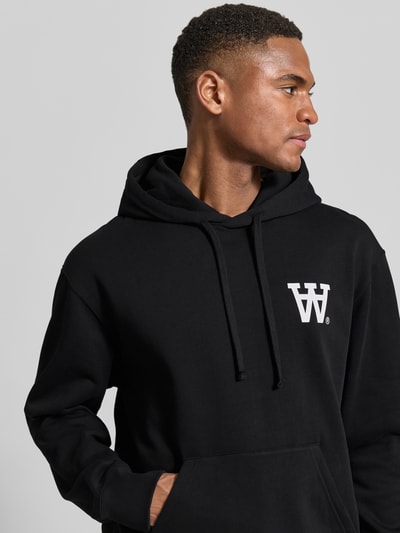 Männermodel trägt schwarzen Hoodie mit Logo-Print und Känguru-Tasche von Wood Wood, lockere Passform, Nahaufnahme Oberkörper.