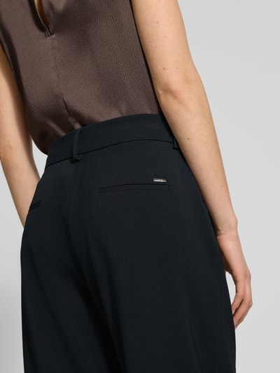 Frauenmodel trägt Wide Fit Stoffhose mit Schurwoll-Anteil Modell 'Megan' von Brax, dunkler Stoff, glatte Textur, Detailaufnahme Rückansicht. Elastischer Material-Mix mit gelegten Falten, eingefassten Beinabschlüssen und Verschlüssen, Innenbeinlänge 68 cm bei Größe 36.