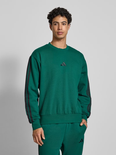Männermodel trägt Sweatshirt mit Logo und Rundhalsausschnitt von adidas Sportswear in Grün, Baumwoll-Viskose-Mix, Relaxed-Fit, Nahaufnahme Oberkörper.