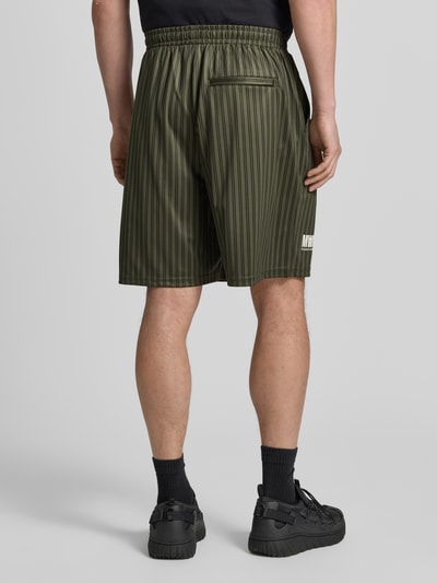 Männermodel trägt Regular Fit Shorts mit Label-Print Modell 'Lama' von Wood Wood in Olivgrün mit schwarzen Streifen, Ganzkörperansicht Rückansicht. Shorts mit elastischem Bund, sichtbarer Label-Print, Bundweite 40 cm, Innenbeinlänge 19 cm.