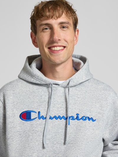 Männermodel trägt grauen Hoodie mit Label-Stitching von Champion aus Baumwoll-Mix, lockerer Schnitt, Nahaufnahme Oberkörper.