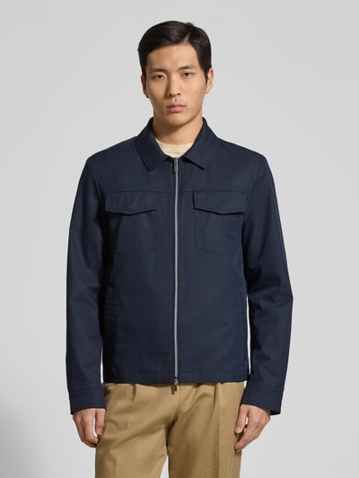 Męski model nosi Jacke mit Pattentaschen und Zweiwege-Reißverschluss od bugatti, bawełniany miks, prosty krój, kolor granatowy, kent kołnierz, zamek dwukierunkowy, kieszenie z patkami i boczne kieszenie, długość tyłu w rozmiarze 50: 67 cm, szerokość ramion w rozmiarze 50: 45 cm, szerokość klatki piersiowej w rozmiarze 50: 57 cm, długość rękawa w rozmiarze 50: 70 cm, pełny widok z przodu