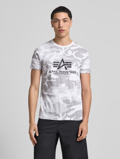 Mężczyznamodel nosi T-Shirt mit Label-Print von Alpha Industries, bawełna, krój regular fit, okrągły dekolt, nadruk z logo, długość tyłu w rozmiarze M: 70 cm, jasnoszary kamuflaż, widok na całą sylwetkę z przodu.