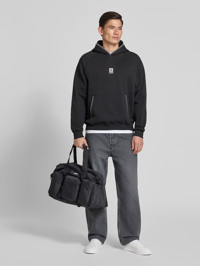 Männermodel trägt Weekender mit Reißverschlusstaschen Modell 'EVERETT' von HUGO, mehrfarbig, Logo Print, Ganzkörperansicht von vorne.