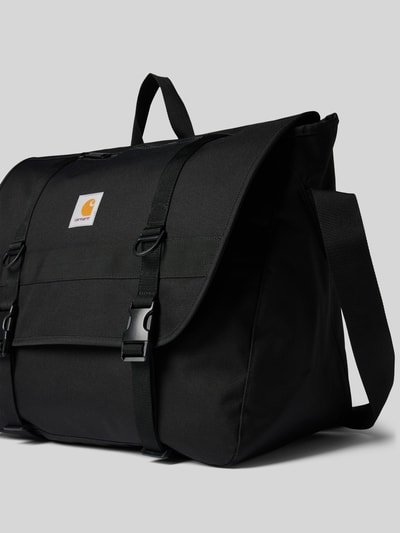 Produktfoto, Messenger Bag mit Label-Patch Modell 'Jake' von Carhartt Work In Progress, schwarze Farbe, robuster Stoff, Hauptfach mit Klickverschluss, Klettverschluss, verstellbarer Schulterriemen, kurzer Henkel, geräumig, dezentes Branding, rechteckige Form, sichtbare Struktur, geeignet für Laptop, neutraler Hintergrund.