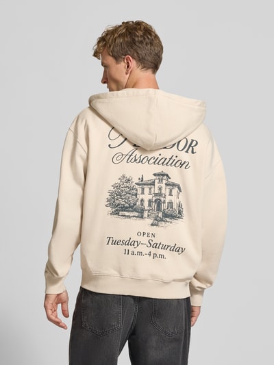Männermodel trägt Relaxed Fit Sweatjacke aus Baumwoll-Mix Modell 'Carloforte' von PEGADOR in Beige mit schwarzem Print, Langarm, gerippte Abschlüsse, überschnittene Schultern, geteilte Kängurutasche, Rückenansicht, Kontext: Herren Sweatjacke, Logo-Stickerei, Maße Größe M.