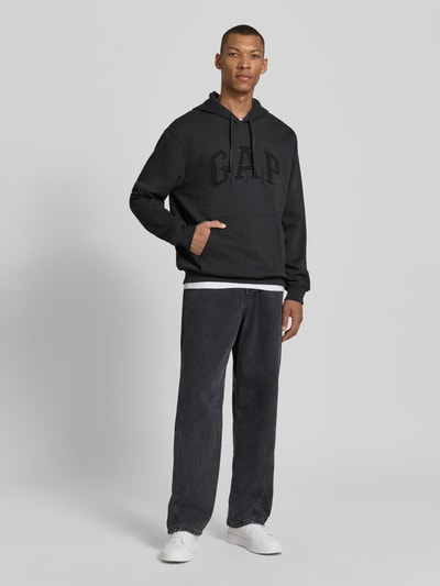 Mannenmodel draagt Hoodie mit Logo-Detail und Känguru-Tasche van GAP in zwart, katoenmix, loose fit, geribde boorden, met capuchon en koord, logo op borst, kangoeroezak; volledige lichaamsaanzicht vooraan.