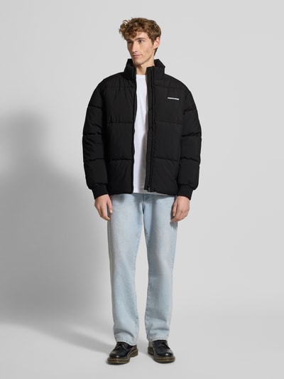 Männermodel trägt schwarze Steppjacke mit Label-Stitching von Pegador, Regular Fit, Stehkragen, Reißverschluss, Ganzkörperansicht von vorne.