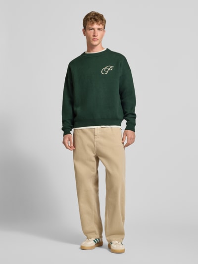 Mannenmodel draagt Relaxed Fit gebreide pullover uit puur katoen van PROHIBITED, labelstitching, ribboorden, losse pasvorm, ruglengte 64 cm, mouwlengte 59 cm, beige broek, groene trui, sneakers, geheellichaamsaanzicht van voren.
