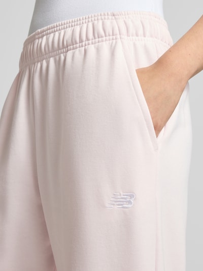 Frauenmodel trägt Sweatpants mit Logo-Stitching von New Balance aus Baumwoll-Mix, Loose-Fit, elastischer Bund, seitliche Eingrifftaschen, elastische Abschlüsse, hellrosa, glatte Oberfläche, Nahaufnahme Oberkörper.