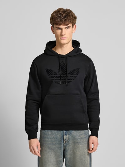 Männermodel trägt Hoodie mit Kapuze von adidas Originals aus Baumwoll-Mix in Schwarz mit Label-Print und Känguru-Tasche, Ganzkörperansicht von vorne.