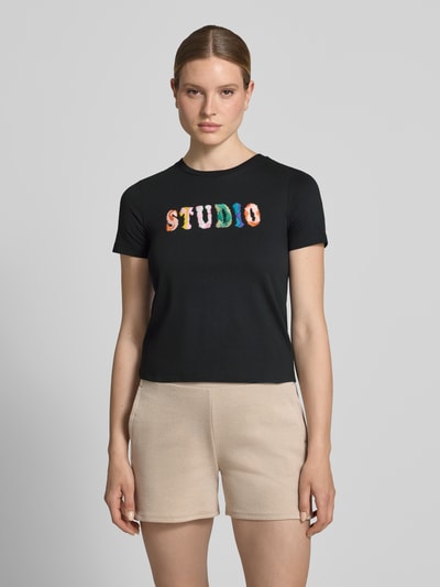 Frauenmodel trägt Regular Fit T-Shirt mit Logo-Stitching von JAKE*S STUDIO WOMAN, schwarze Baumwolle, beiger Shorts, Rundhalsausschnitt, Ganzkörperansicht von vorne.