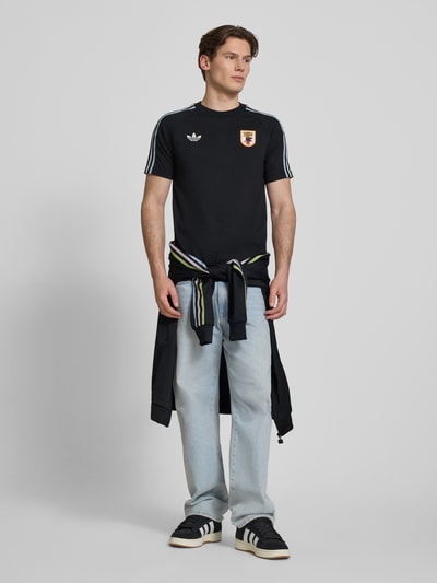 Männermodel trägt Regular Fit T-Shirt aus Baumwoll-Mix mit JFA-Applikation Modell 'NOIR' von adidas Originals, Optik: Unifarben, Logo-Stitching, Kontraststreifen, Rundhals, Ärmelform: Kurzarm, Herren T-Shirt, Ganzkörperansicht von vorne.