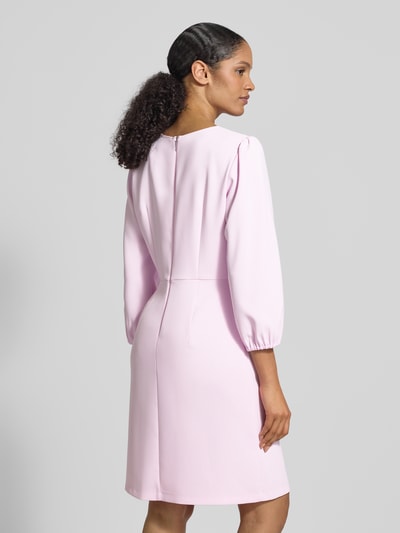 Modelka nosi Knielanges Kleid mit Rundhalsausschnitt und 3/4-Arm od Marc Cain, jasnoróżowy kolor, materiał z domieszką stretchu, prosty krój, długość do kolan, szerokość w rozmiarze 36: 43 cm, długość tyłu: 95 cm, długość rękawa: 53 cm, widok całej sylwetki z tyłu.