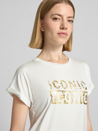 Frauenmodel trägt T-Shirt mit Rundhalsausschnitt und Print Modell 'MARICA' von Soyaconcept, Weiß mit goldfarbenem Schriftzug, Lyocell-Elasthan-Mix, Ärmel mit Aufschlag, Nahaufnahme Oberkörper.