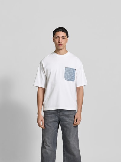 Männermodel trägt Loose Fit T-Shirt aus reiner Baumwolle von Dsquared2 in Weiß mit Denim-Brusttasche und Nieten, Ganzkörperansicht von vorne.