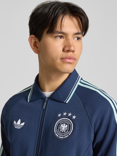 Männermodel trägt Regular Fit Trainingsjacke mit Deutschland Logo von adidas Sportswear, dunkelblau, weiße Kontraststreifen, Nahaufnahme Oberkörper