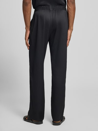 Męski model nosi czarne spodnie z plisą, loose fit, Hose mit elastischem Bund Modell 'Houston' von Les Deux, szerokość w rozmiarze M 42 cm, długość nogawki 81 cm, tekstura gładka, całkowity widok z tyłu.
