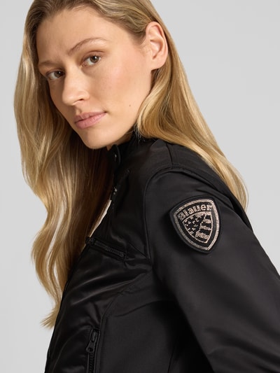 Frauenmodel trägt Jacke mit Zweiwege-Reißverschluss Modell 'MELBA' von Blauer Usa, schwarze Farbe, glatte Textur, Nahaufnahme Oberkörper. Körpernahe Passform, Stehkragen mit Druckknopf, Reißverschlusstaschen und Label-Patch sichtbar.