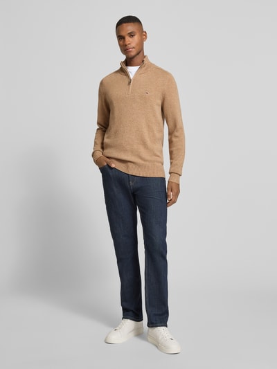 Męski model nosi Regular Fit Troyer aus reiner Wolle od Tommy Hilfiger, dzianinowy sweter beżowy, logo, krój regular fit, długo rękaw, widok całej sylwetki z przodu.