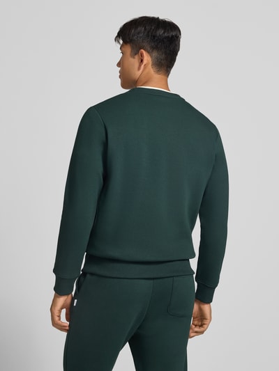 Männermodel trägt Regular Fit Sweatshirt mit Baumwoll-Anteil Modell 'Munk' von JACK & JONES in Dunkelgrün, glatte Textur, Ganzkörperansicht Rückansicht. Material-Mix mit Baumwoll-Anteil, elastische Abschlüsse, Rundhalsausschnitt, Rückenlänge 60 cm, Ärmellänge 67 cm.