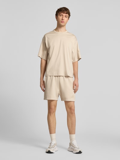 Męski model nosi Regular Fit Sweatshorts mit Modal und Baumwoll-Anteil od adidas Sportswear, efekt: jednolity kolor, krój regular fit, detal z logo, elastyczny ściągacz z tunelem, wpuszczane kieszenie, mieszanka materiałów z domieszką modalu i bawełny, jasnobeżowy kolor, gładka tekstura, całkowity widok z przodu.