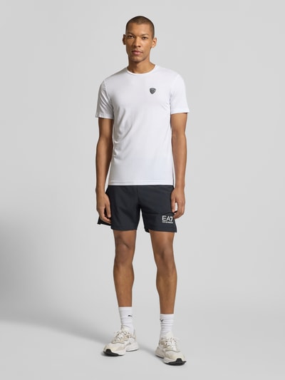 Männermodel trägt T-Shirt aus Viskose-Mix mit Label-Patch von EA7 Emporio Armani in Weiß, Herren T-Shirt, Slim-Fit, Ganzkörperansicht von vorne.