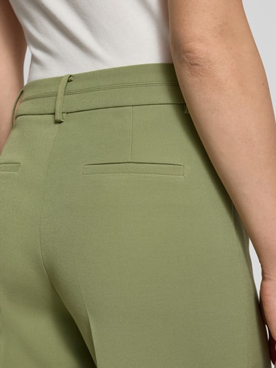 Frauenmodel trägt Stoffhose mit Bundfalten von More & More in hellem Grün, glatte Textur, Gesäßtaschen sichtbar, Nahaufnahme Rückansicht. Material-Mix mit Stretch-Anteil, lockerer Schnitt, Detailansicht der Gesäßtaschen und Bund.