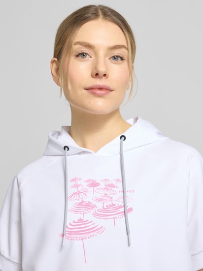 Modelka kobieca ma na sobie T-Shirt aus Baumwoll-Mix mit Kapuze Modell 'SHARON3' od FIRE + ICE, biały kolor, różowy nadruk, krótki rękaw, mieszanka bawełny i elastanu, luźny krój, tunel w kapturze i u dołu, elastyczne zakończenia rękawów, detal z logo, widok z przodu, zbliżenie na górną część ciała.