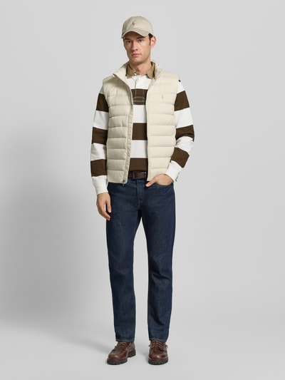 Männermodel trägt Regular Fit Weste in Stepp-Optik von Polo Ralph Lauren, cremefarben, mit Stehkragen, Reißverschluss, Logo-Stitching, Eingrifftaschen, gefüttert, dazu gestreiftes Oberteil und Jeans; Rückenlänge 69 cm, Schulterbreite 44 cm, Brustweite 58 cm, Ganzkörperansicht von vorne.