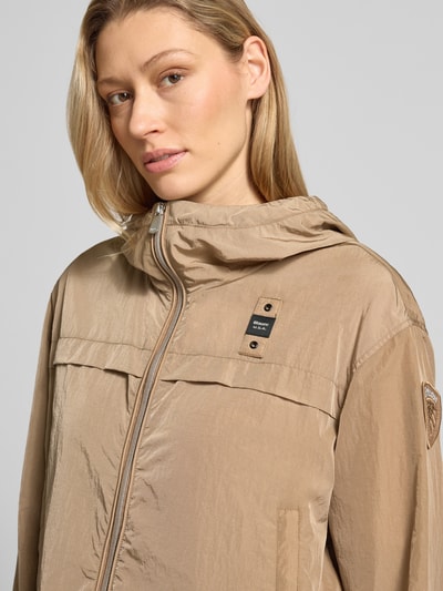 Frauenmodel trägt beige Trekking Jacke mit Kapuze Modell 'BURRAGE' von Blauer Usa, glatte Textur, Nahaufnahme Oberkörper. Gerader Schnitt, Zweiwege-Reißverschluss, Label-Patches, seitliche Reißverschlusstaschen, elastische Ärmelabschlüsse, Rückenlänge 56 cm, Schulterbreite 48 cm, Brustweite 56 cm, Ärmellänge 57 cm bei Größe S.