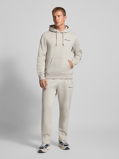 Mężczyzna model nosi Straight Fit Sweatpants mit Label-Stitching od CHAMPION, kolor jasny, mieszanka bawełny, elastyczny pas, haftowane logo, długość wewnętrzna nogawki 70 cm, widok całej sylwetki z przodu.