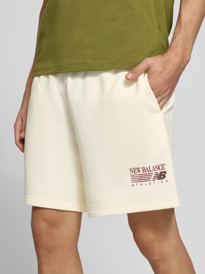 Männermodel trägt Sweatshorts aus Baumwoll-Mix von New Balance in Creme mit Label-Print, elastischem Bund, Nahaufnahme Oberschenkel. Innenbeinlänge 20 cm, Bundweite 41 cm, gerade Passform.