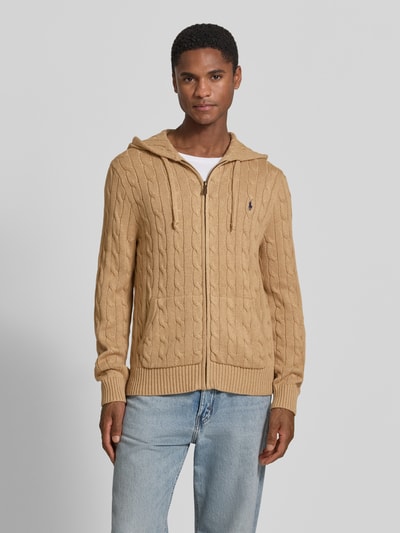 Męski model nosi Regular Fit Strickjacke aus reiner Baumwolle von Polo Ralph Lauren, beżowy kardigan rozpinany, wzór w warkocze, gładka tekstura, Ganzkörperansicht von vorne.