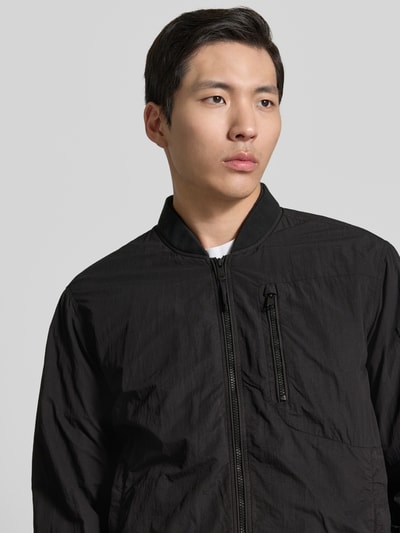 Männermodel trägt schwarzen Blouson mit Label-Patches Modell 'IBE' von khujo, Reißverschluss, Brusttasche, Nahaufnahme Oberkörper.