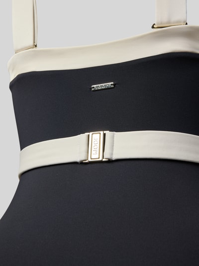 Frauenmodel trägt Badeanzug mit breiten Trägern Modell 'Blanca Beach' von JOOP! Collection in Schwarz mit cremefarbenem Taillenband, Nahaufnahme Oberkörper.