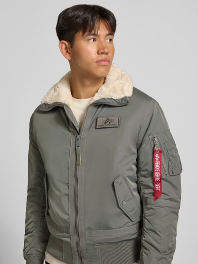 Männermodel trägt Bomberjacke mit Label-Patch und Kentkragen von Alpha Industries, olivgrün, Teddyfellkragen, Nahaufnahme Oberkörper. Rückenlänge 65 cm, Schulterbreite 45 cm, Brustweite 56 cm, Ärmellänge 65 cm.