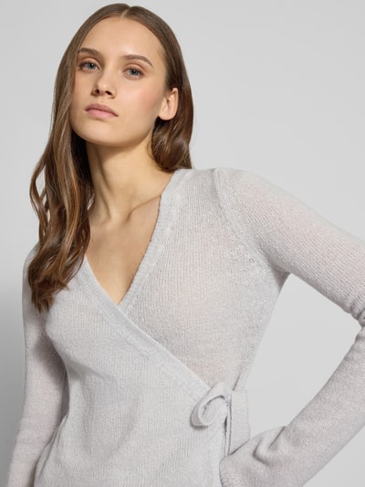 Frauenmodel trägt Strick-Cardigan in Wickeloptik von Review, hellgrau, V-Ausschnitt, lange Ärmel, Schleifenverschluss, Nahaufnahme Oberkörper.