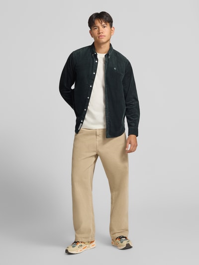 Männermodel trägt Regular Fit Freizeithemd mit Logo-Stitching Modell 'MADISON' von Carhartt Work In Progress, Ganzkörperansicht von vorne, reine Baumwolle, Button-Down-Kragen, Knopfleiste, Sportmanschetten, abgerundeter Saum, beige Hose, dunkelgrünes Hemd
