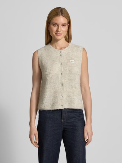Modelka nosi Kamizelkę w imitacji dzianiny od American Vintage, mieszanka materiałów z domieszką wełny z alpaki, krój regular fit, prążkowane wykończenia, okrągły dekolt, listwa guzikowa, detal z logo, długość tyłu w rozmiarze S: 49 cm, jasnobeżowy, struktura puszysta, widok całej sylwetki z przodu.