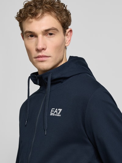 Männermodel trägt Slim Fit Sweatjacke mit Kapuze von EA7 Emporio Armani in Dunkelblau mit Logo-Print, Nahaufnahme Oberkörper.