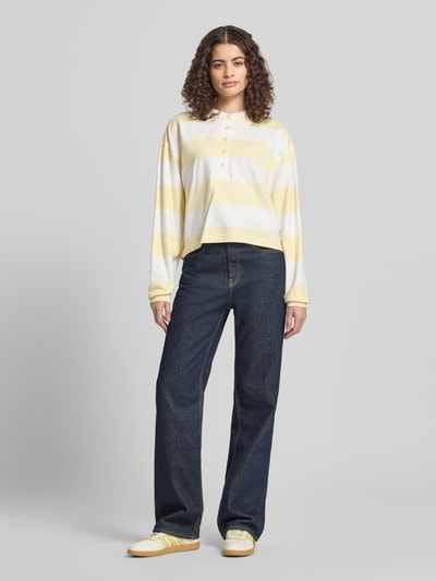 Frauenmodel trägt Relaxed Fit Sweatshirt mit Logo-Stitching Modell 'Varsity' von Calvin Klein Jeans, weiß-gelbe Streifen, reine Baumwolle, elastische Ärmelabschlüsse, Knopfleiste, langärmeliges Design, kombiniert mit dunkelblauer Hose, Ganzkörperansicht von vorne.