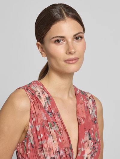 Kobieta nosi Regular Fit Maxikleid mit floralem Allover-Print od ADLYSH, kwiatowy nadruk na całej powierzchni, dekolt w serek, krój bez rękawów, obszycie falbanami, wycięcia, wiązanie, plisy, kolor różowy, blady róż i kremowy, materiał lekko połyskujący, szerokość w rozmiarze S: 32 cm, długość tyłu w rozmiarze S: 52 cm. Zbliżenie na górną część ciała, ujęcie z przodu.