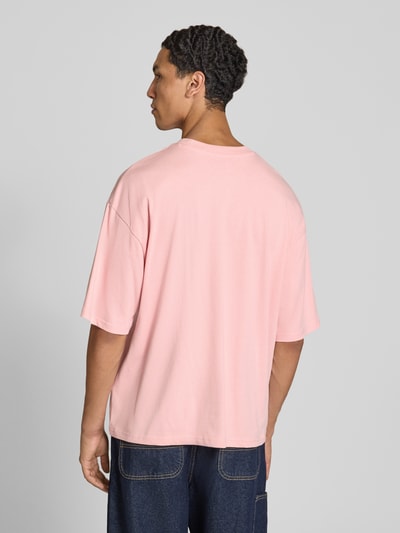 Męski model nosi Oversized T-Shirt aus reiner Baumwolle od REVIEW, kolor jasnoróżowy, krój oversized, okrągły dekolt, nadruk z logo, długość tyłu w rozmiarze M: 66 cm, widok całej sylwetki z tyłu.