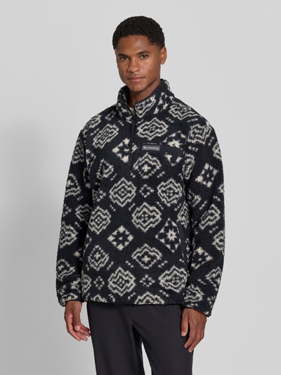Męski model ma na sobie Regular Fit Pullover mit Logo-Patch od Columbia, Polar, krój regular fit, naszywka z logo, kołnierz zapinany, Długość tyłu w rozmiarze M: 70 cm, Długość rękawów w rozmiarze M: 68 cm, czarno-biała faktura, wzór geometryczny, widok całej sylwetki od przodu.