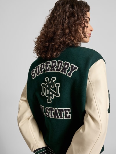 Frauenmodel trägt College Jacke mit Motiv- und Logo-Stitchings von Superdry im Woll-Mix, Lockerer-Schnitt, Stehkragen, Druckknopfverschluss, Rückenlänge 62 cm, Schulterbreite 49 cm, Brustweite 60 cm, Ärmellänge 60 cm, elastische Abschlüsse, Ganzkörperansicht Rückansicht.