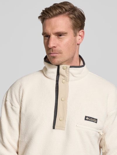 Męski model nosi Fleecepullover mit Stehkragen und Knopfleiste od Columbia, krój regular fit, wyhaftowane logo, kieszeń zapinana na zamek błyskawiczny, kremowa faktura, zbliżenie na górną część ciała